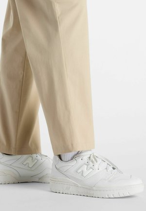 Baskets en cuir blanc avec des accents texturés, design à lacets et logo "N" bien en vue. Associées à un pantalon beige et des chaussettes blanches.