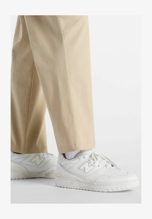 Zapatillas blancas de cuero con acentos texturizados, diseño de cordones y prominente logo "N". Combinadas con pantalones beige y calcetines blancos.