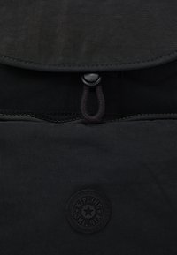 Black fabric bag with a rounded flap, button closure, and a circular embossed logo at the bottom center. Smooth texture and minimalistic design.