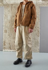 Brun bomulds zip-up jakke med hætte, beige løse cargobukser og sorte snørebåndsstøvler. Jakken har et logo patch på brystet.