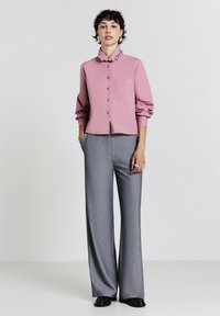 Donna che indossa una camicia rosa con bottoni e collare di perline, pantaloni larghi grigi, scarpe nere e orecchini d'argento, in piedi con le mani nelle tasche.