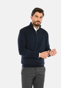 Maglione blu navy con zip e collo a coste, realizzato in un morbido tessuto a maglia, abbinato a pantaloni grigi.