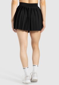 Zwarte geplooide rok met een elastische tailleband, met een gladde textuur en gelaagd ontwerp. G gedragen met witte sportschoenen en sokken.