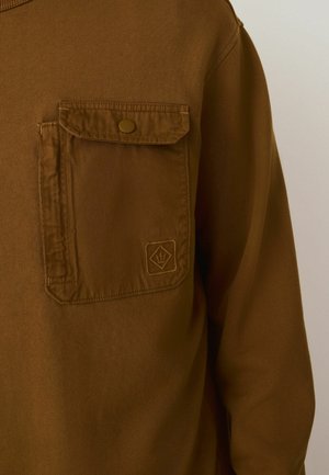 Brown long-sleeve shirt with a textured fabric, featuring a chest pocket with a snap closure and a logo patch in the bottom corner.
