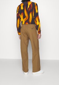 Dlouhý rukáv košile s motivem plamenů v oranžové a černé barvě, spárovaná s hnědými kalhotami s uvolněným střihem a dvěma zadními kapsami.