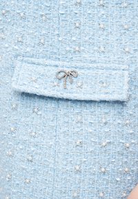 Υφασμα από ανοιχτό μπλε τουίντ με μικρές διακοσμήσεις από πέρλες και ασημένιες χάντρες, διαθέτει τσέπη με καπάκι και μικρό μπιζού φιόγκο με στρας.