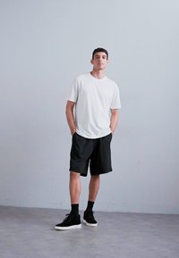 Hvit t-skjorte, sorte shortser og svarte joggesko. Modellen står mot en ensfarget grå bakgrunn, med hendene i lommene, iført en avslappet passform.