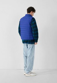 Mann, der eine blaue gesteppte Weste über einem dunkelgrünen und blauen karierten Hemd, hellblauen Jeans und weißen Sneakern trägt und abgewandt steht.