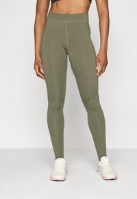 Leggings ajustados en color verde oliva con una cinturilla alta, textura suave y costuras en contraste. Combinados con zapatillas deportivas de color claro.