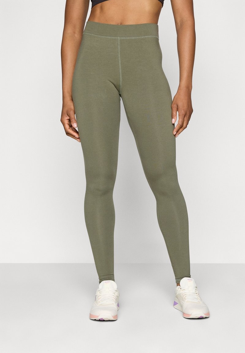 Leggings ajustados en color verde oliva con una cinturilla alta, textura suave y costuras en contraste. Combinados con zapatillas deportivas de color claro.
