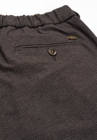 Pantalon à carreaux gris foncé avec taille élastique, poche arrière boutonnée et petite étiquette de marque en métal près de la poche.