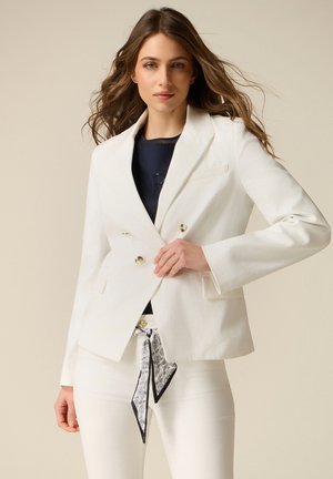 Kvinde iført en hvid dobbeltradet blazer over en sort top, hvide bukser med et mønstret tørklæde bundet om taljen, stående mod en beige baggrund.