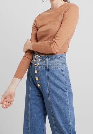 Kobieta w brązowej, prążkowanej bluzce z długim rękawem, włożonej do wysokich, szerokich spodni dżinsowych w kolorze niebieskim, z szerokim paskiem i złotymi guzikami.