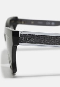 Occhiali da sole neri con montatura in plastica elegante e stanghette trasparenti caratterizzate da un motivo testurizzato e un dettaglio con logo. Gli accenti valorizzano il design.