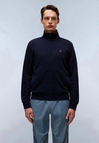 Jovem vestido com uma jaqueta azul-marinho com fecho éclair, com um pequeno logotipo vermelho no peito e calças azul-claro, de pé contra um fundo claro e neutro.