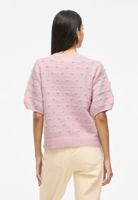 Suéter rosa de punto con patrones de relieve en forma de pompones, mangas cortas abullonadas y dobladillo acanalado. Combinado con pantalones beige, que muestran un ajuste relajado.