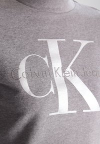 Šedá bavlněná mikina s velkým bílým logem "CK" a textem "Calvin Klein Jeans", s jemnou texturou a klasickým kulatým výstřihem.