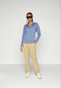 Giacca in pile azzurra chiara con chiusura a zip, maniche lunghe e tasche laterali, abbinata a pantaloni beige e sneaker di colore chiaro.
