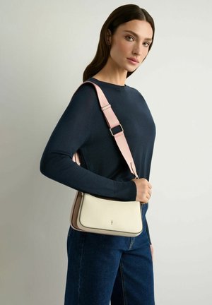 Kobieta w ciemnoniebieskim swetrze i dżinsach niesie kremową torebkę na szerokim różowym pasku na ramieniu na jednolitym tle.