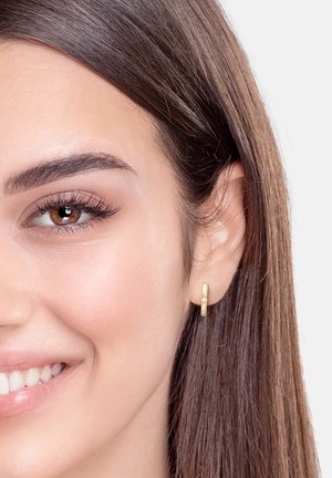 Goldene Creolenohrringe mit einem glatten, abgerundeten Design und einem kleinen Akzent am Rand. Das Model hat langes, glattes braunes Haar.