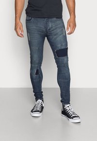 Mörkblå slitna skinny jeans med revor och lagade sektioner. Bärs med svarta hög-top sneakers med vita snören.