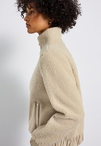Giacca in pile beige con colletto alto, tasche laterali e orlo elasticizzato. Caratterizzata da un tessuto morbido e testurizzato e da un colore neutro.