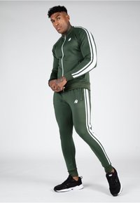 Groene trainingsoutfit met witte strepen, bestaande uit een jack met rits en aansluitende broek. Bevat een klein logo op de borst en op de broek. Zwarte sneakers.