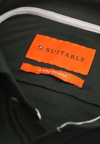 Donkergroen stof met een herringbone structuur, voorzien van oranje labels met de tekst "GESCHIKT" en "4 Richtingen Rek" op de stof gestikt.