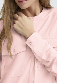 Camicia button-up rosa chiaro con due tasche frontali, maniche lunghe e bottoni ai polsini. Tessuto morbido e design dal taglio rilassato.