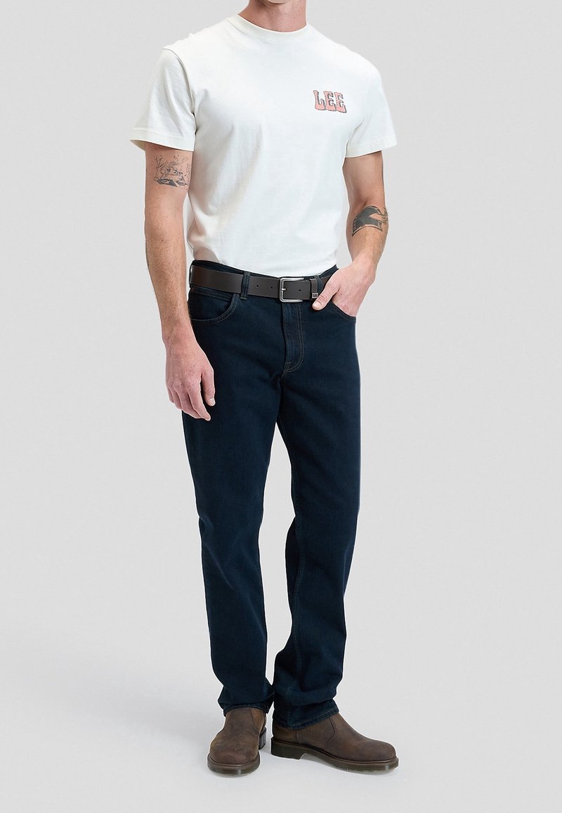 Homme portant un t-shirt blanc Lee, une ceinture noire, un jean bleu foncé et des bottes marron, debout avec une main dans la poche devant un fond uni.