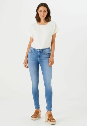 Weißes kurzärmliges Oberteil mit Rundhalsausschnitt, kombiniert mit hellblauen Skinny-Jeans und braunen Plateausandalen mit geflochtenem Fußbett.