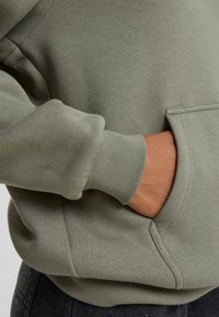 Sweat à capuche vert olive avec poignets côtelés, poche avant et texture douce en polaire, conçu pour le confort et la chaleur.