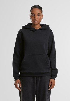 Femme aux cheveux foncés tirés en arrière portant un sweat à capuche noir et un pantalon noir, debout devant un fond gris uni.