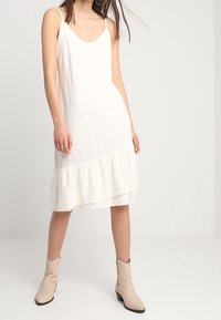 Vit ärmlös klänning med rundad halsringning och volangkant. Bärs med beige cowboyboots, som har en slät textur och en flödande design.