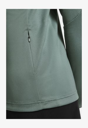 Zbliżenie na zielony, teksturowany top z długim rękawem z kieszenią na suwak po boku i widocznymi detalami szwów.