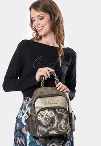 Frau in einem schwarzen Oberteil und einem bedruckten Rock, die eine gemusterte Handtasche mit geflochtenen Griffen und goldfarbenen Akzenten hält, lächelt und schaut nach unten.