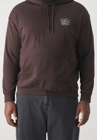 Uomo che indossa un felpa con cappuccio marrone scuro con un piccolo sole bianco e un faccina sorridente sul petto e pantaloni grigio scuro.
