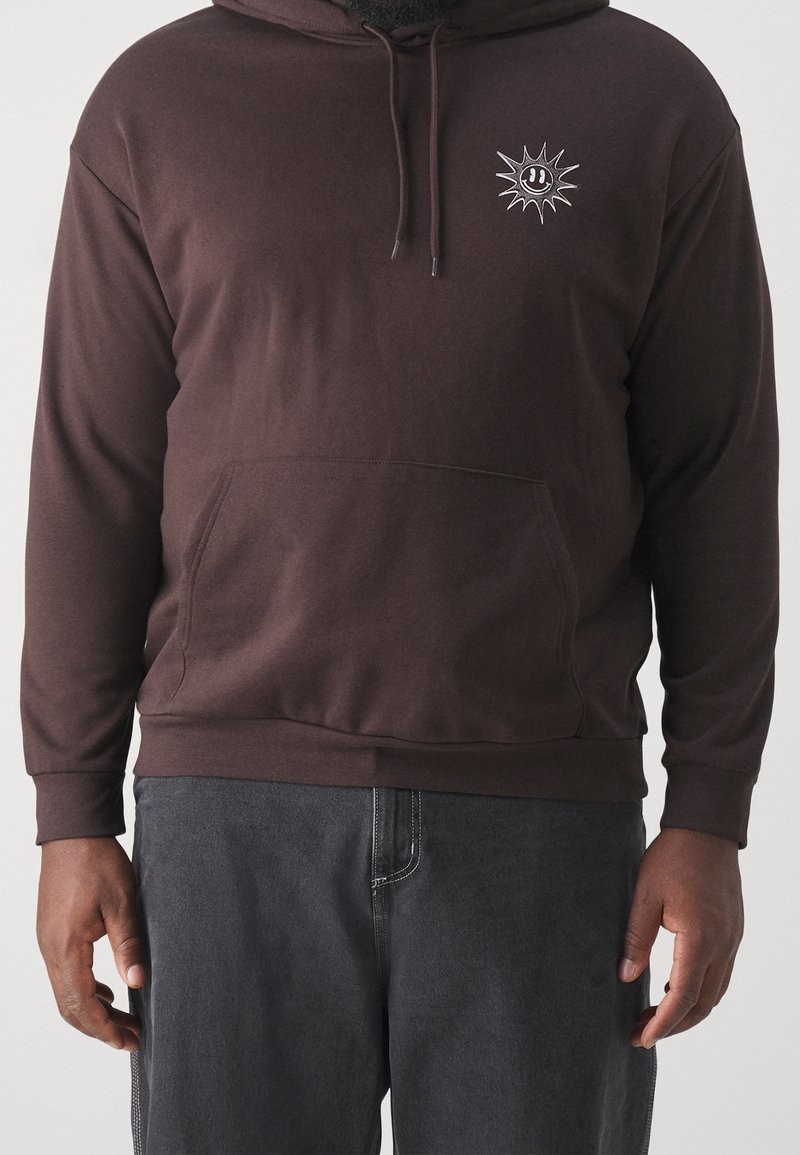Uomo che indossa un felpa con cappuccio marrone scuro con un piccolo sole bianco e un faccina sorridente sul petto e pantaloni grigio scuro.