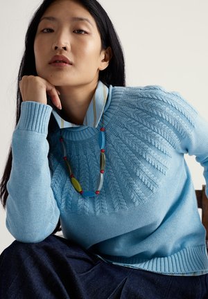 Suéter de punto azul claro con diseño texturizado alrededor del escote, llevado sobre una camisa de rayas con cuello. Se ve tela vaquera oscura debajo.