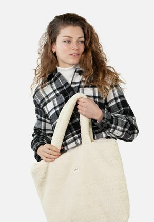 Tote bag in cream plush material with a soft texture. Features wide handles and a simple tag, held by a person in a black and white plaid shirt.