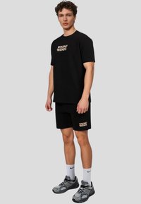 Zwarte katoenen t-shirt en short set met de gouden tekst "WRONG FRIENDS". Model draagt grijze sneakers en witte crew-sokken. Casual fit, korte mouwen.
