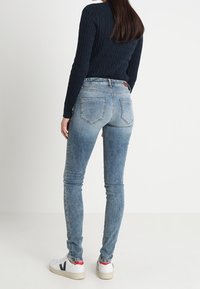 Kvinna klädd i ljusblå skinny jeans, marinblå kabelstickad tröja och vita sneakers med röd och svart logotyp, som står mot en vit bakgrund.