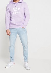 Fialová mikina s kapucí s bílým logem Adidas, kombinovaná se světle modrými džínami a bílými teniskami, s ohrnutými manžetami a tkaničkami.