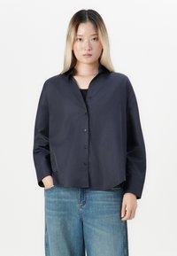 Woman with straight blonde hair wearing a loose dark button-up shirt and wide-leg blue jeans, standing against a white background.