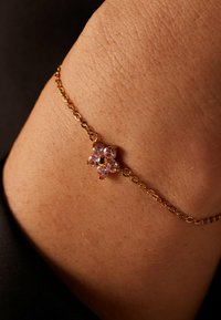 Gouden armband met een bloemencharme, versierd met roze edelstenen en een centrale gouden accent. Close-up op de huid die de ingewikkelde details toont.