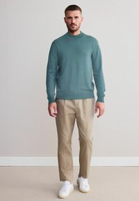 Lyse teal strikket sweater, beige bukser og hvide sneakers. Enkel, tætsiddende design med lange ærmer og rund halsudskæring.