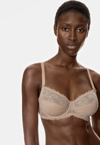 Soutien-gorge en dentelle beige clair, présentant de délicats motifs floraux, des bretelles réglables et une finition à volants le long des bords.