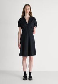 Robe polo noire à manches courtes, avec un décolleté en V et une coupe décontractée. Tissu doux, atteignant le mi-cuisse. Portée avec des chaussettes et des chaussures noires.
