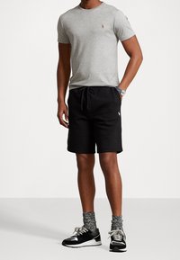 Man i grå kortärmad t-shirt, svarta shorts med dragsko, grå strumpor, svart och vit sneakers samt en klocka mot en enkel bakgrund.