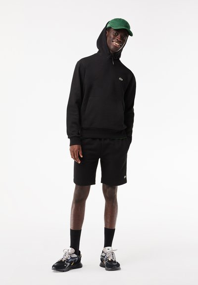 Hoodie en coton noir avec une poche kangourou, associé à un short noir. Logo vert remarquable sur la poitrine et casquette verte. Baskets texturées.